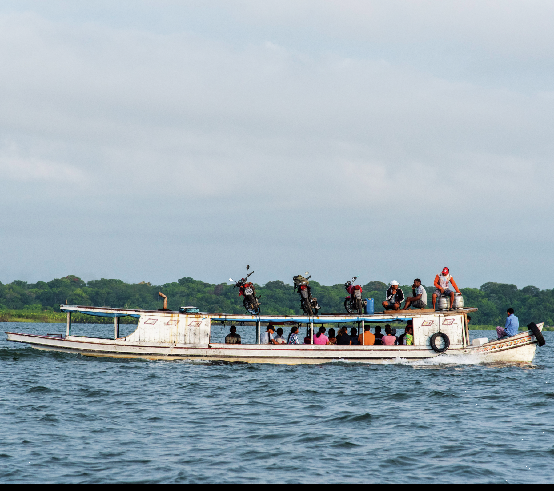 Fotografia. No rio, barco branco transportando várias pessoas de diferentes etnias. Na parte superior do barco, há pessoas e motocicletas. Ao fundo, árvores e céu azul com nuvens.