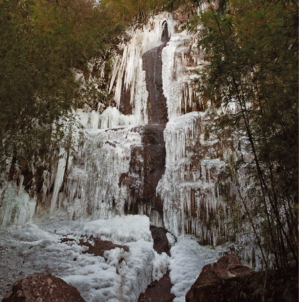 Fotografia. Cachoeira congelada. A formação rochosa tem vários níveis e a queda de água está congelada. Ao redor, algumas árvores.