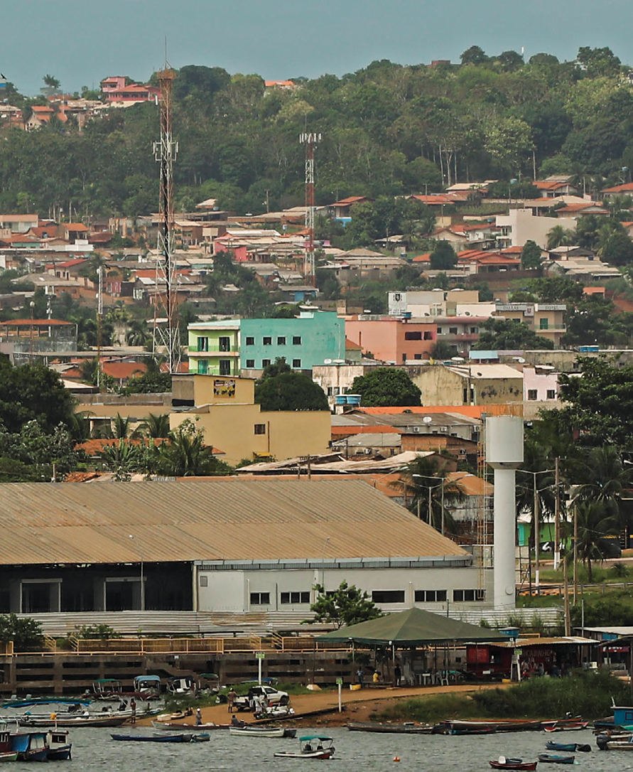 Fotografia. Cidade de Altamira. Na parte inferior da foto, margem do rio Xingu na cor cinza, com embarcações de diferentes tamanhos e cores. Acima da margem, uma construção com telhado marrom. Ao fundo, diferentes construções, com tamanhos e cores diversas, com estruturas semelhantes a casas, e antenas espalhadas. Mais ao fundo, na parte superior da imagem, vegetação com árvores e céu azul. Atrás da vegetação, são visíveis algumas casas.