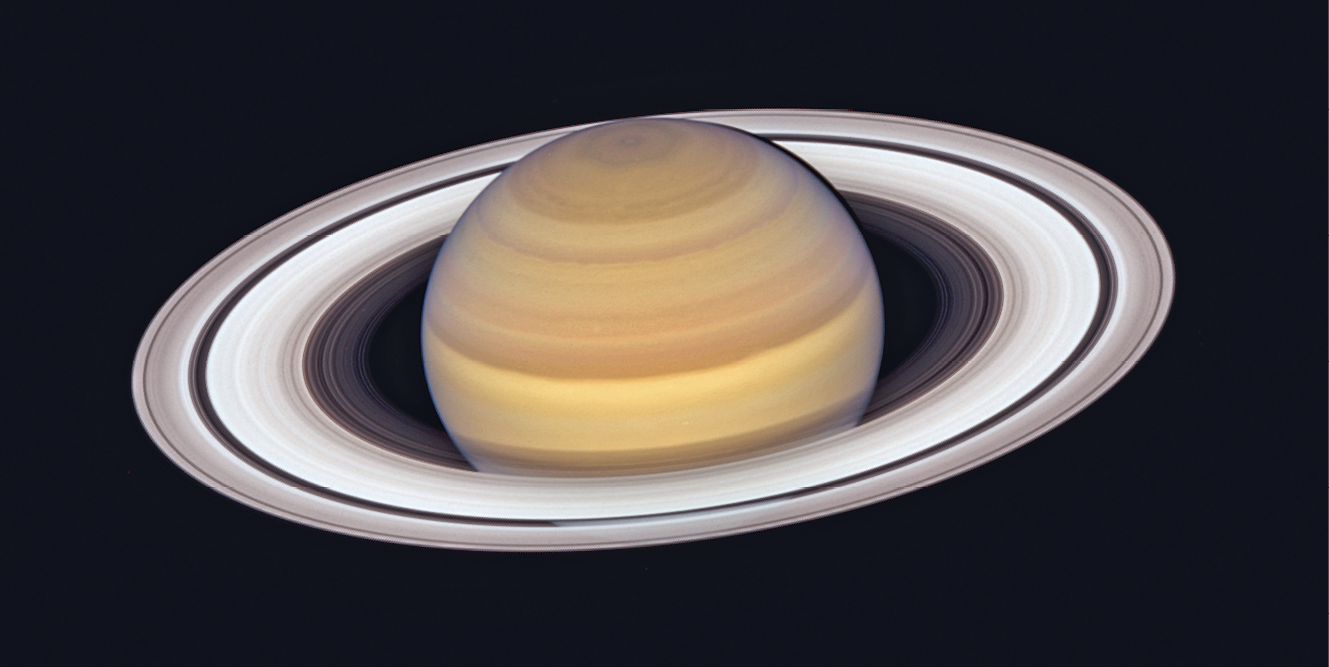 Fotografia. Planeta Saturno. Em fundo preto, no centro da imagem, esfera com faixas em tons de bege e marrom. Em volta da esfera, anéis em tons de branco e cinza.