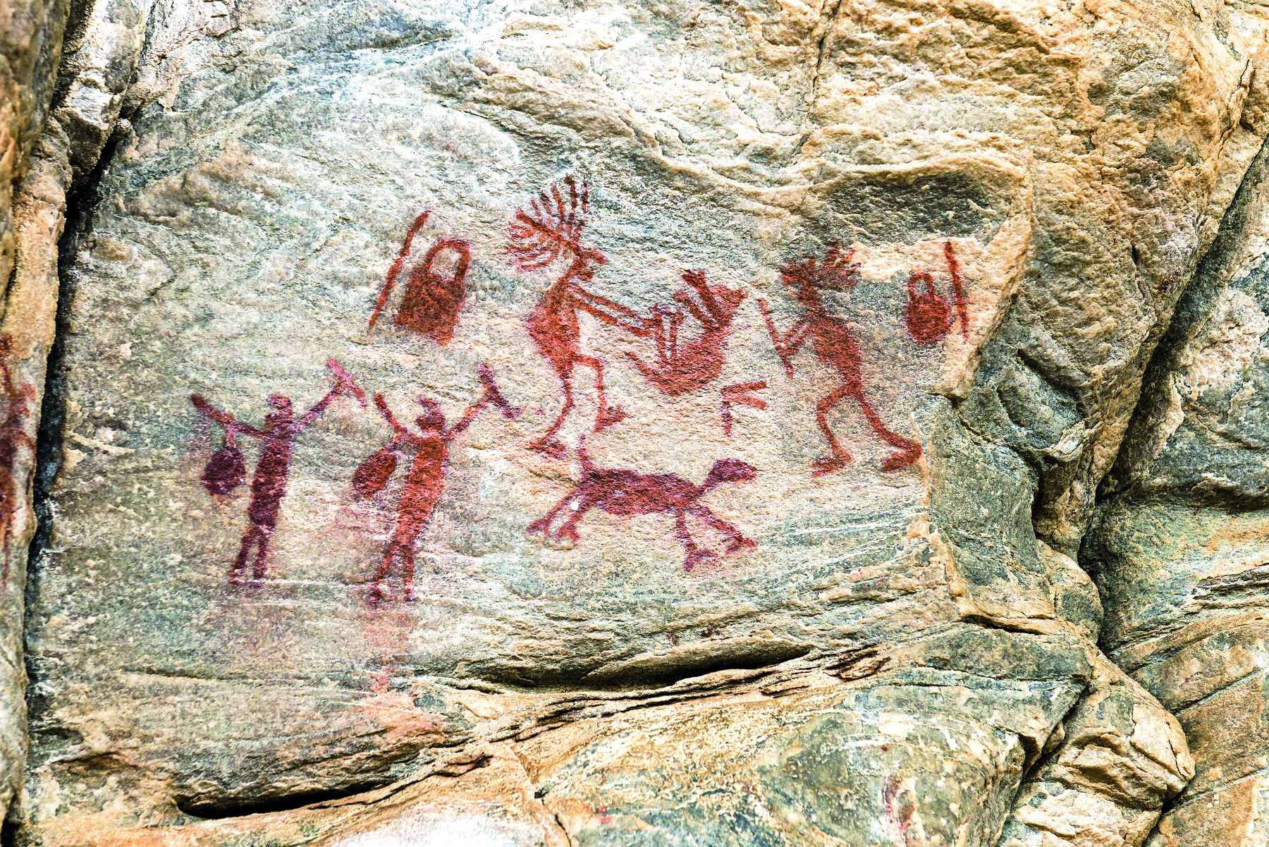 Fotografia. Pintura rupestre em uma parede de rocha. Na pintura, feita com tinta vermelha, há silhuetas de duas pessoas segurando objetos e duas caçando um animal, que está com as patas para cima. Ao lado delas há outros objetos e um animal quadrúpede.