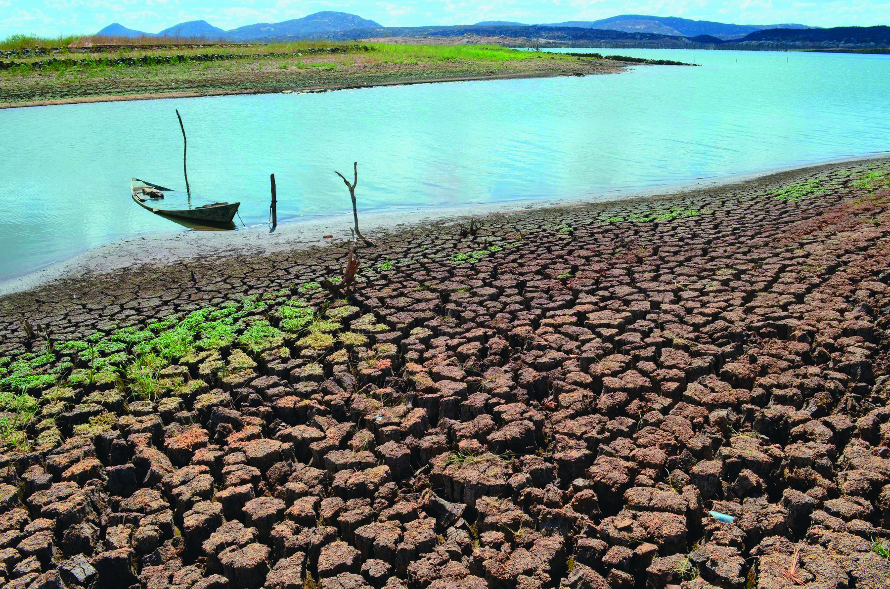 Fotografia. Imagem colorida, formato paisagem. Barragem, um rio com um pequeno barco amarrado a um tronco. Ao redor, terra seca.