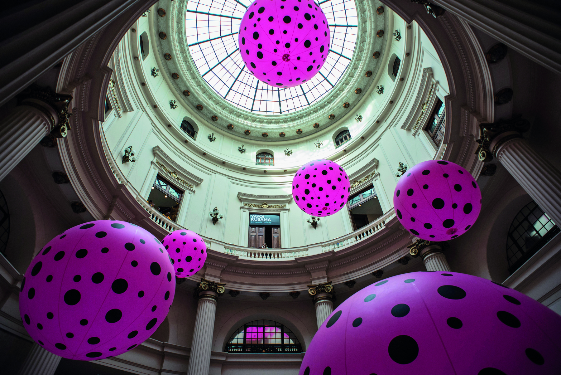 Fotografia. Vista de baixo para cima da parte interna de um edifício antigo. No alto há uma abóbada circular de vidro e, mais abaixo, diversas janelas, portas e pilares. Ocupando o ambiente, há enormes balões cor-de-rosa com bolinhas pretas suspensos.