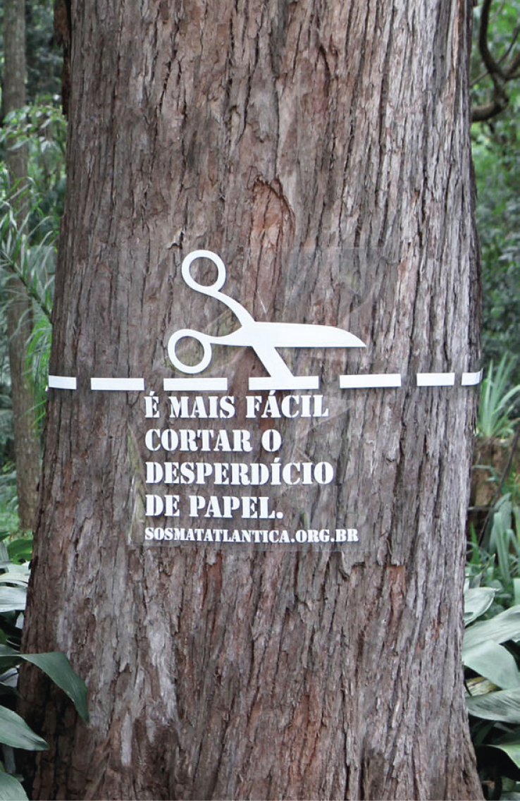 Cartaz. A imagem mostra a fotografia de um tronco de árvore. Sobreposto ao tronco, há o desenho de uma linha branca pontilhada, dividindo o tronco ao meio. Acima da linha, uma tesoura branca em movimento de corte. Abaixo da linha, o texto: É MAIS FÁCIL CORTAR O DESPERDÍCIO DE PAPEL.