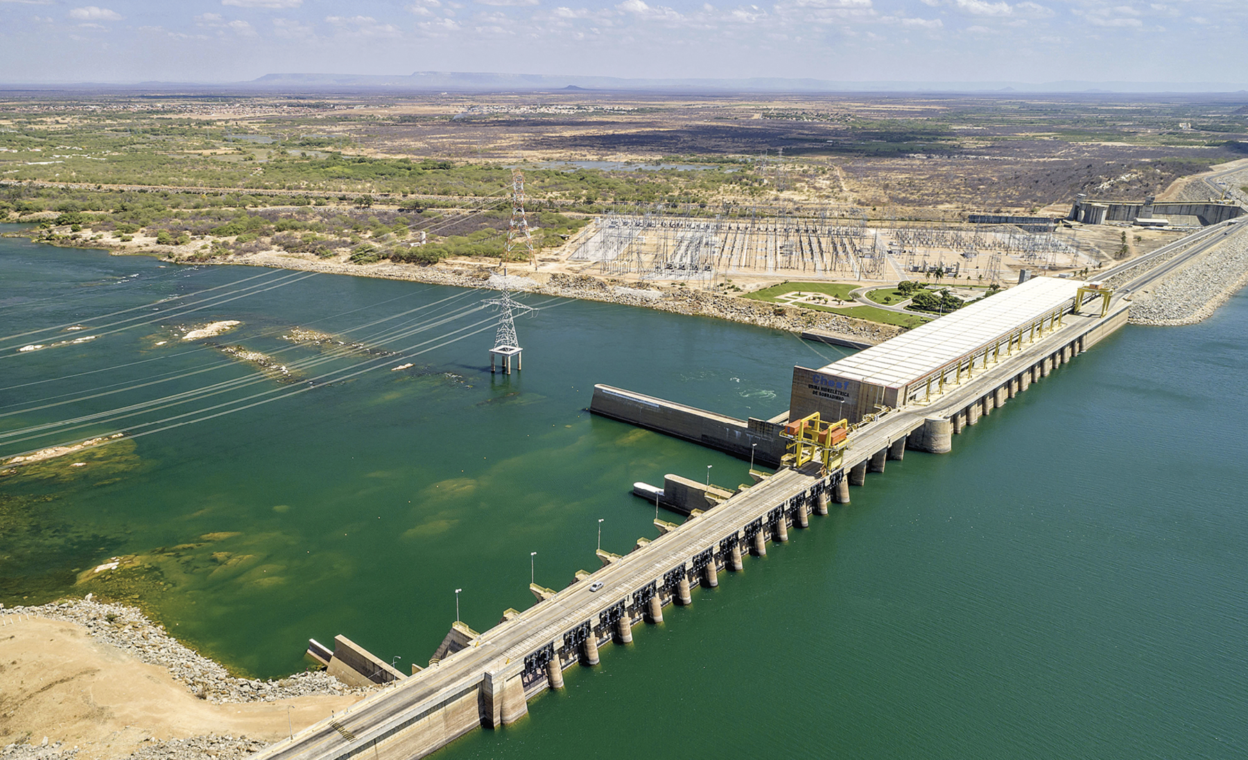 Fotografia. Imagem colorida, formato paisagem. Vista aérea de uma barragem composta por rio, cabos de energia elétrica e uma ponte com aberturas sobre a água.  No fundo, vegetação.