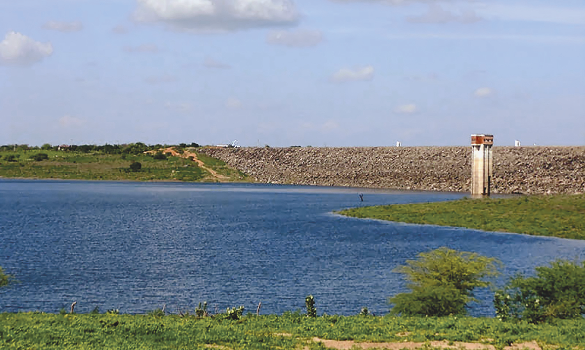 Fotografia. Imagem colorida, formato paisagem. Uma barragem de água. Ao redor, vegetação.  Acima, céu com nuvens.