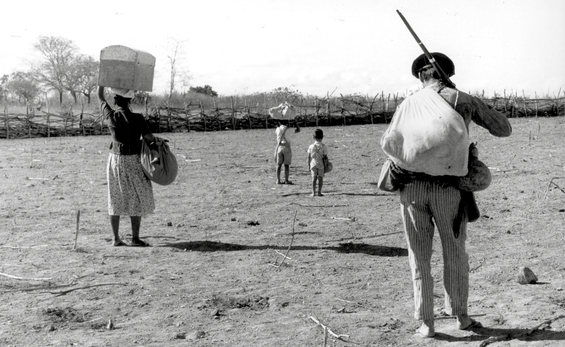 Fotografia. Imagem em preto e branco. Formato paisagem. A foto mostra quatro pessoas, dois adultos e duas crianças, caminhando em um terreno árido. À esquerda da foto, uma mulher usa blusa e saia. Ela está de costas, segurando uma caixa sobre a cabeça e uma trouxa no braço direito. À direita, um homem de chapéu, blusa e calça listrada. Ele está de costas, com uma trouxa nas costas e segurando uma espingarda. À frente deles, duas crianças vestidas com camisa e shorts. Elas estão descalças e segurando trouxas.