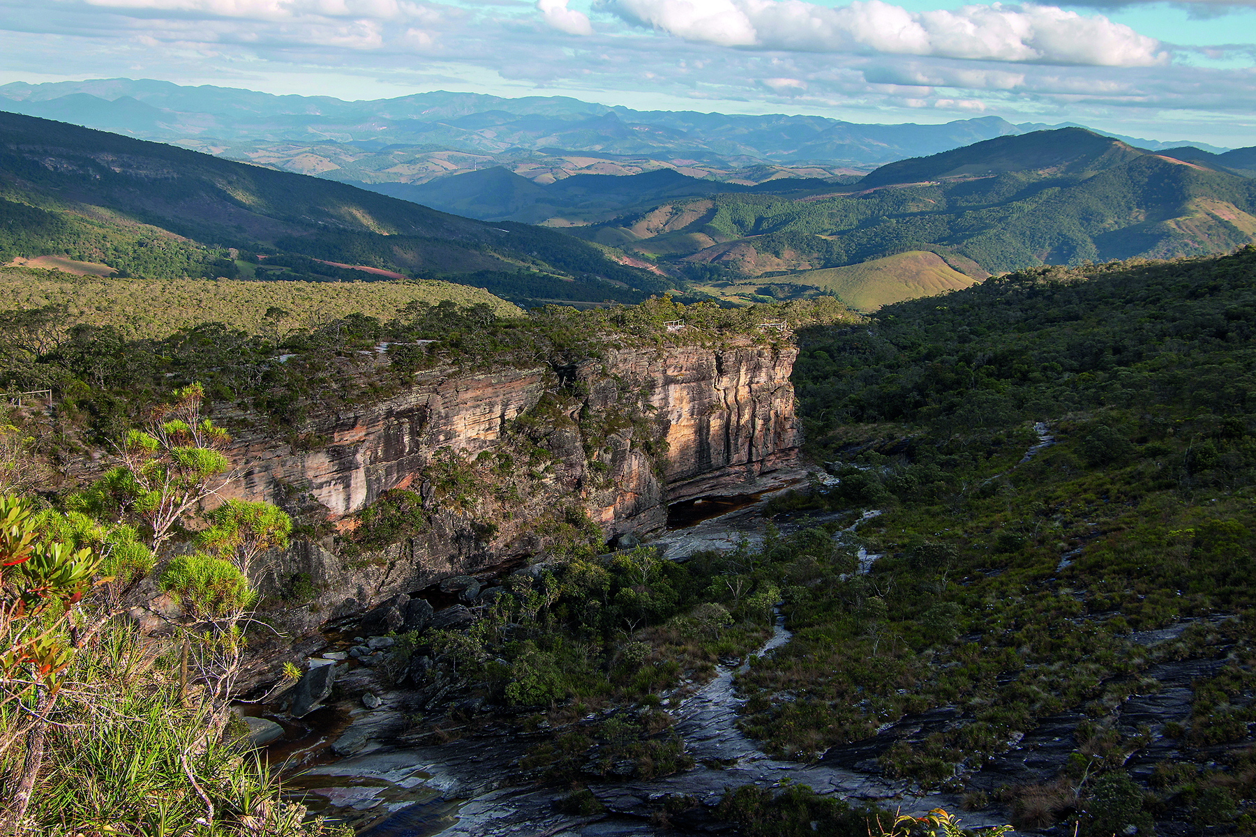 Fotografia. Paisagem natural montanhosa que se estende até a linha do horizonte e totalmente coberta por vegetação, sob céu nublado. À frente da imagem, um aparente trecho de relevo erodido percorrido pelo leito de um curso de água.