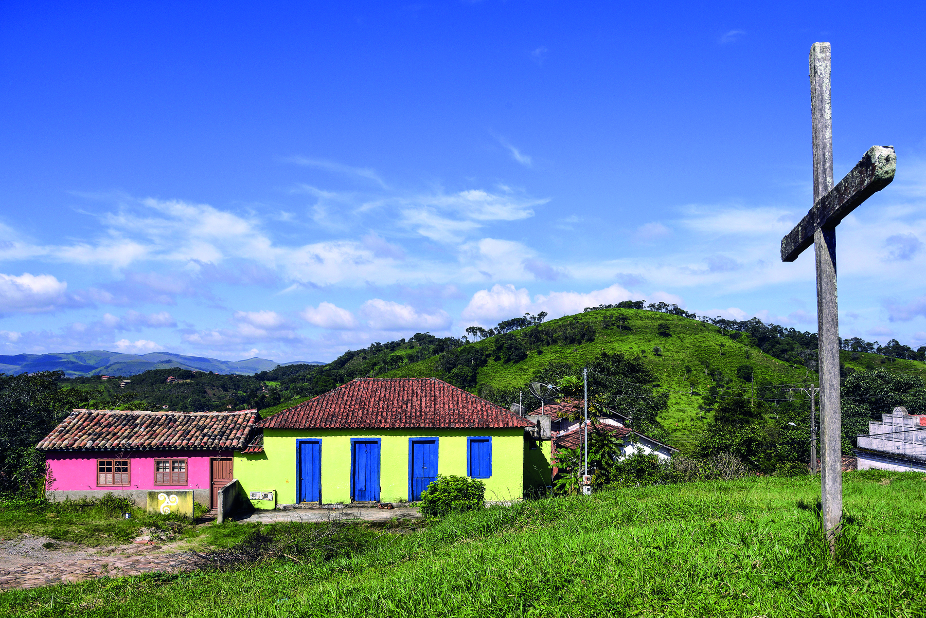 Fotografia. À esquerda, duas casas pequenas de fachadas coloridas de rosa, amarelo e azul em uma área rural, junto a um morro, em terreno coberto de vegetação. À direita, há uma cruz de madeira fixada em um gramado. Ao fundo, paisagem montanhosa sob céu azul parcialmente nublado.