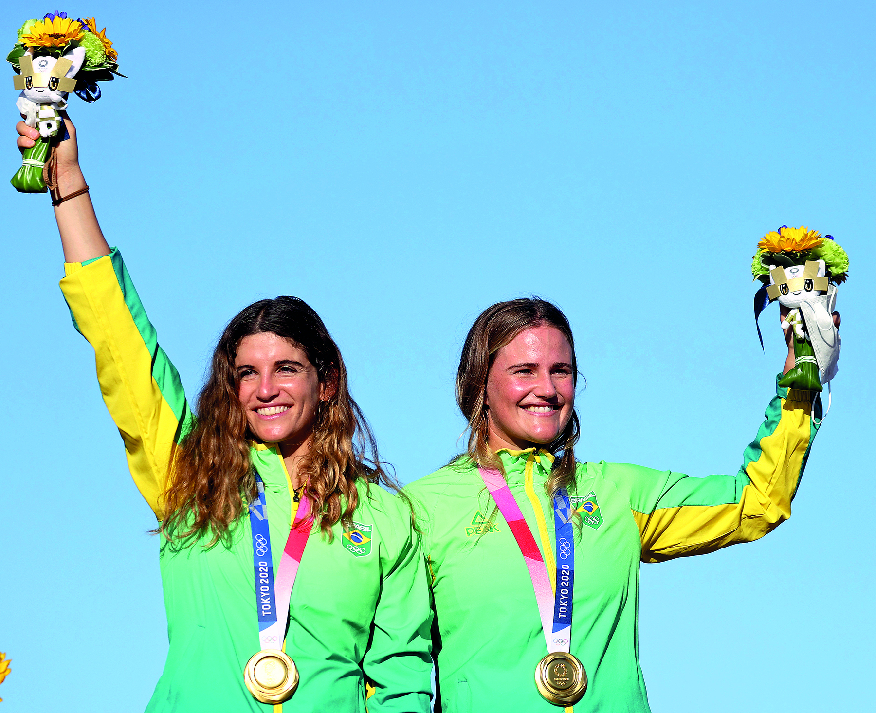 Fotografia. Retrato de duas atletas lado a lado. À esquerda, uma mulher com cabelos longos ondulados, usando uniforme do Brasil e medalha no pescoço. Ela está com o braço direito esticado para cima, segurando um buquê de flores. À direita, uma mulher com cabelos presos para trás, usando uniforme do Brasil e medalha no pescoço. Ela está com o braço esquerdo semiflexionado para cima, segurando um buquê de flores. Elas estão sorrindo.