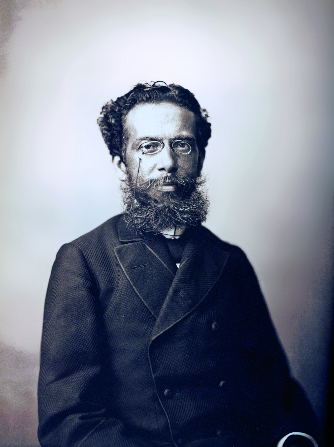 Fotografia em preto e branco. Retrato de um homem com cabelos curtos e barba grande, usando óculos e terno.