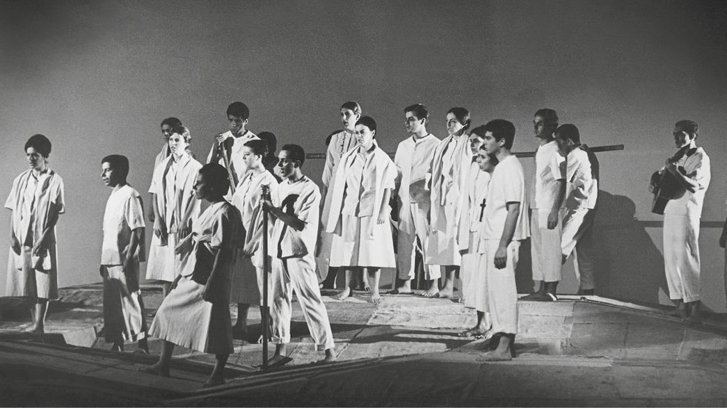 Fotografia em preto e branco. Um grupo de pessoas em cena teatral. Elas estão de pé, voltadas para a esquerda. Usam roupas claras e estão descalças. Um homem está atrás de todo o grupo e segura um instrumento musical de corda.