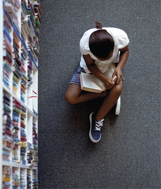 Fotografia. Vista de cima, uma jovem sentada no chão, com as pernas cruzadas, vestindo camiseta, short e tênis. Ela está com as mãos sobre um livro aberto, que está apoiado nas suas pernas. Sua cabeça está voltada para baixo, na direção do livro. Do lado esquerdo, estantes com livros.