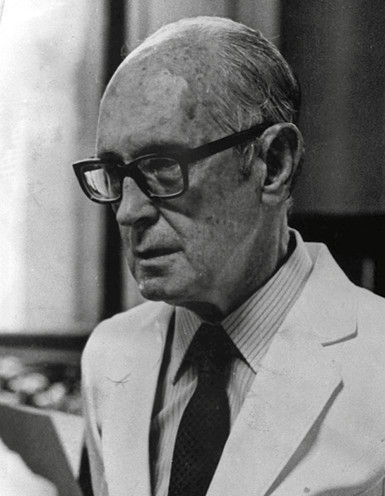 Fotografia em preto e branco. Retrato em meio perfil de um homem calvo, de óculos, rosto enrugado, vestindo terno claro, camisa e gravata.