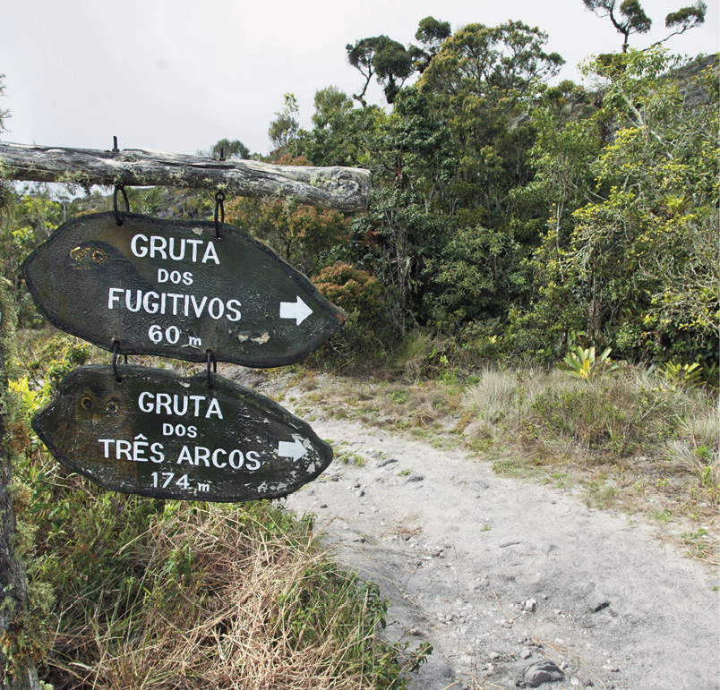 Fotografia. À esquerda, duas placas de troncos de árvore suspensos em um galho, com os dizeres pintados em letras e números brancos: GRUTA DOS FUGITIVOS 60 m e GRUTA DOS TRÊS ARCOS 174 m. À direita, uma trilha em meio a área rural cercada de vegetação com arbustos.