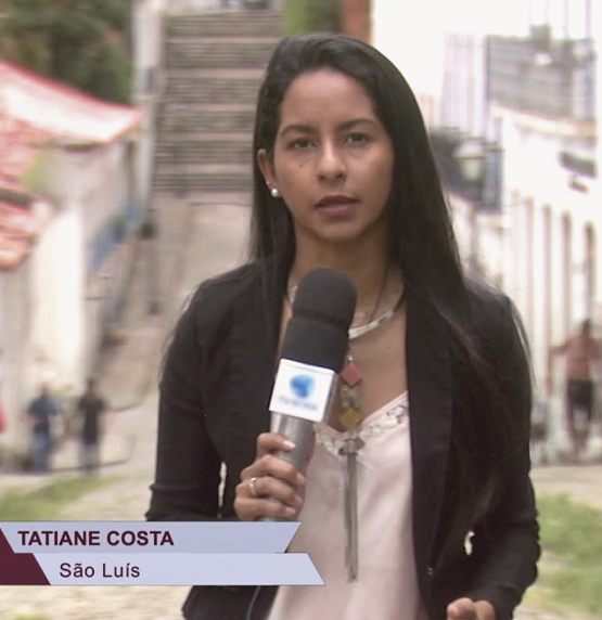 Fotografia. Mulher de cabelos castanhos vestindo blusa de mangas longas, usando brinco e colar, está em pé de frente e segura um microfone com a mão direita. Ao fundo, cenário urbano com casas e edifícios.