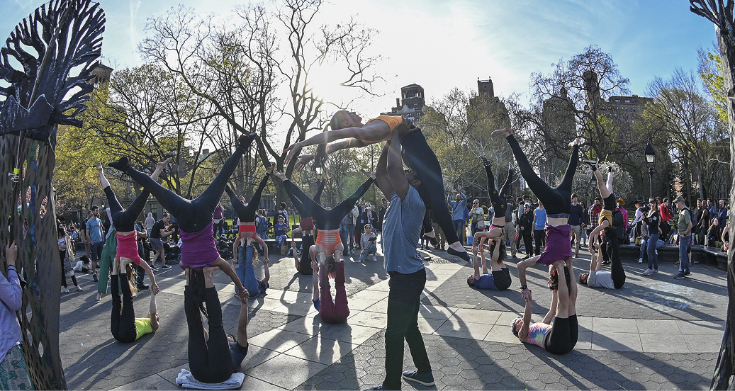 Fotografia. Manifestação artística a céu aberto. Grupo de jovens vestindo roupas coloridas pratica em duplas movimentos de equilibrismo semelhantes aos de espetáculos circenses: alguns se apoiam deitados de costas no chão, com as pernas estendidas para cima, enquanto o colega com quem cada um deles forma dupla se equilibra de cabeça para baixo, com as pernas afastadas estendidas para cima, enquanto apoia os ombros nos pés do colega que está deitado, segurando as mãos deste. Alguns estão em pé de braços erguidos, sobre os quais se apoia o colega de dupla, que estende braços e pernas suspensos no ar, arqueando o corpo para trás. Ao redor, pessoas observando os jovens. Ao fundo, área arborizada sob céu azul com algumas nuvens brancas.