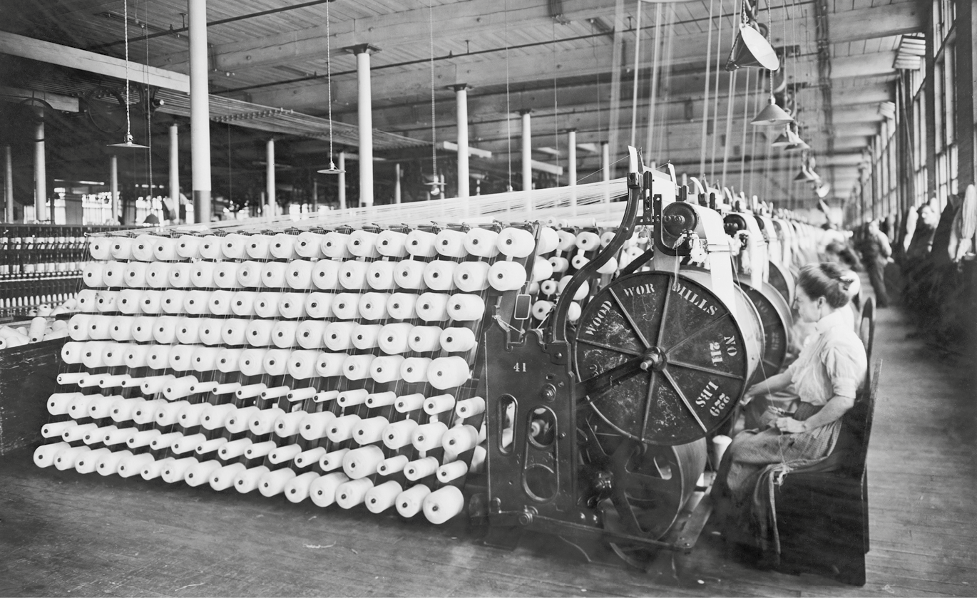 Fotografia em preto e branco. Ambiente industrial. Do lado esquerdo, inúmeras bobinas de linhas enfileiradas. Do lado direito, mulheres de perfil sentadas operando máquinas têxteis dispostas lado a lado em uma fila que se estende por todo o comprimento do galpão. Do alto, fios de linha que abastecem as máquinas.