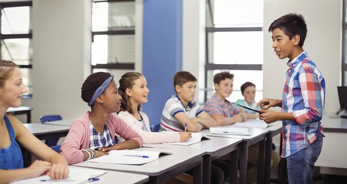 Fotografia. Turma de estudantes em uma sala de aula. À esquerda, seis jovens estão sentados em carteiras dispostas lado a lado. Todos estão olhando na direção de um jovem, à direita da imagem, que está em pé, vestindo camisa xadrez e calças jeans, com um tablet em uma das mãos, fazendo uma apresentação. Ao fundo, janelas envidraçadas permitem a entrada da luz natural no ambiente.