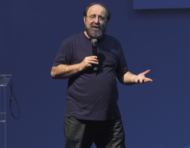 Fotografia. Homem meio calvo de óculos, de cabelos, barba e bigode grisalhos vestindo camisa azul e calça jeans. Ele está com o braço direito semiflexionado à altura do peito e segura um microfone sem fio, posicionando-o próximo do rosto, e seu braço esquerdo está semiflexionado para a frente, com a mão espalmada para cima. Ao fundo, cor azul-escura.