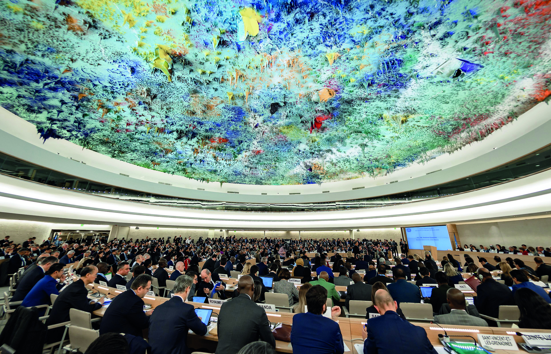 Fotografia. Uma grande sala em formato circular com muitas pessoas reunidas em torno de uma apresentação. Na parte inferior, dezenas de pessoas de costas usando terno e roupa social. A maioria delas está sentada à mesa. As cadeiras e as mesas estão organizadas em fileiras circulares. Há sobre a mesa telas e papéis. No fundo da sala, uma tela exibe uma apresentação. Ao lado da tela, algumas pessoas estão em pé. Na parte superior, teto circular azul claro com relevos coloridos.