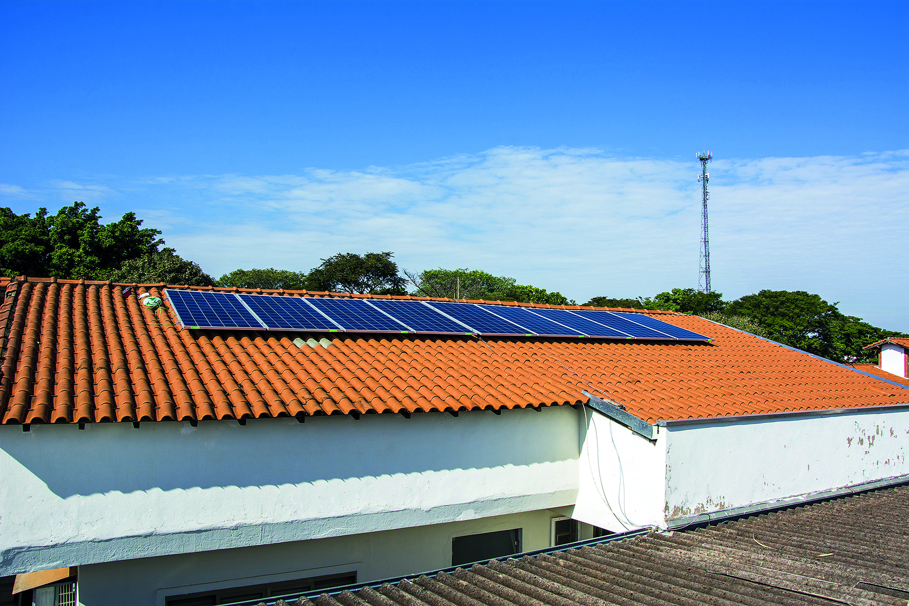 Fotografia. Painéis de captação da energia solar dispostos sobre o telhado de uma construção. Ao fundo copas de árvores e uma torre de telecomunicações sob céu azul com algumas nuvens.