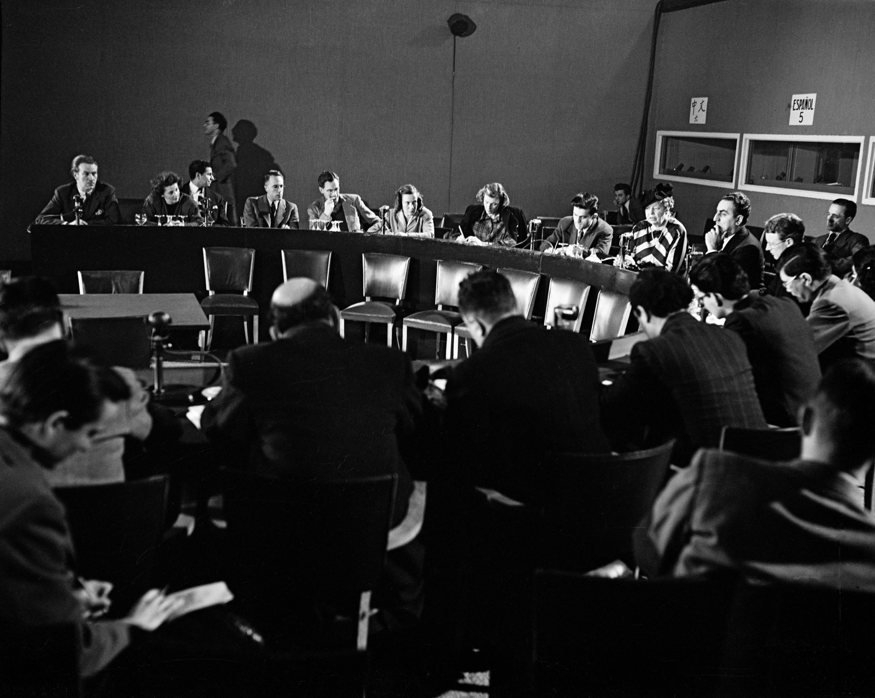 Fotografia em preto e branco. Grupo de pessoas sentadas em torno de uma bancada oval.