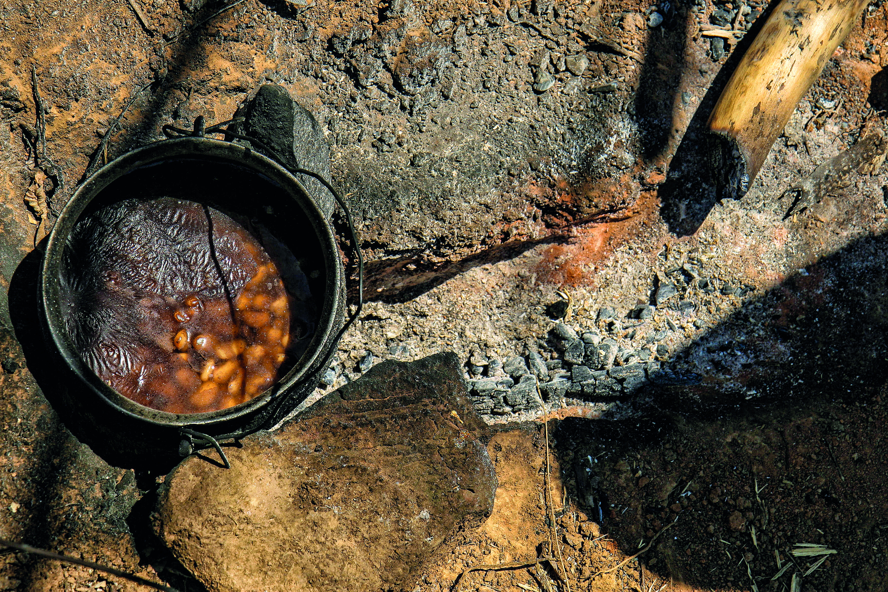 Fotografia. Uma panela de material escurecido com alguns grãos no interior está apoiada em algumas pedras, ao lado de uma tora de lenha chamuscada em uma extremidade.