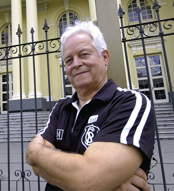 Fotografia. Retrato de um homem com cabelos brancos, usando camiseta esportiva preta com detalhes em branco. Atrás, fachada de uma construção com portão de grades.