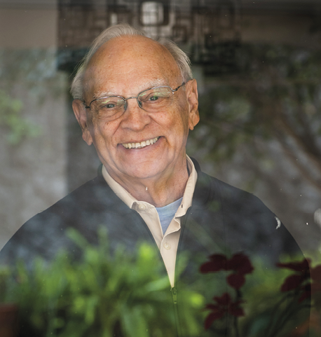 Fotografia. Retrato frontal de um homem sorridente, meio calvo e de cabelos brancos, de óculos, vestindo camiseta azul, camisa bege e jaqueta preta.