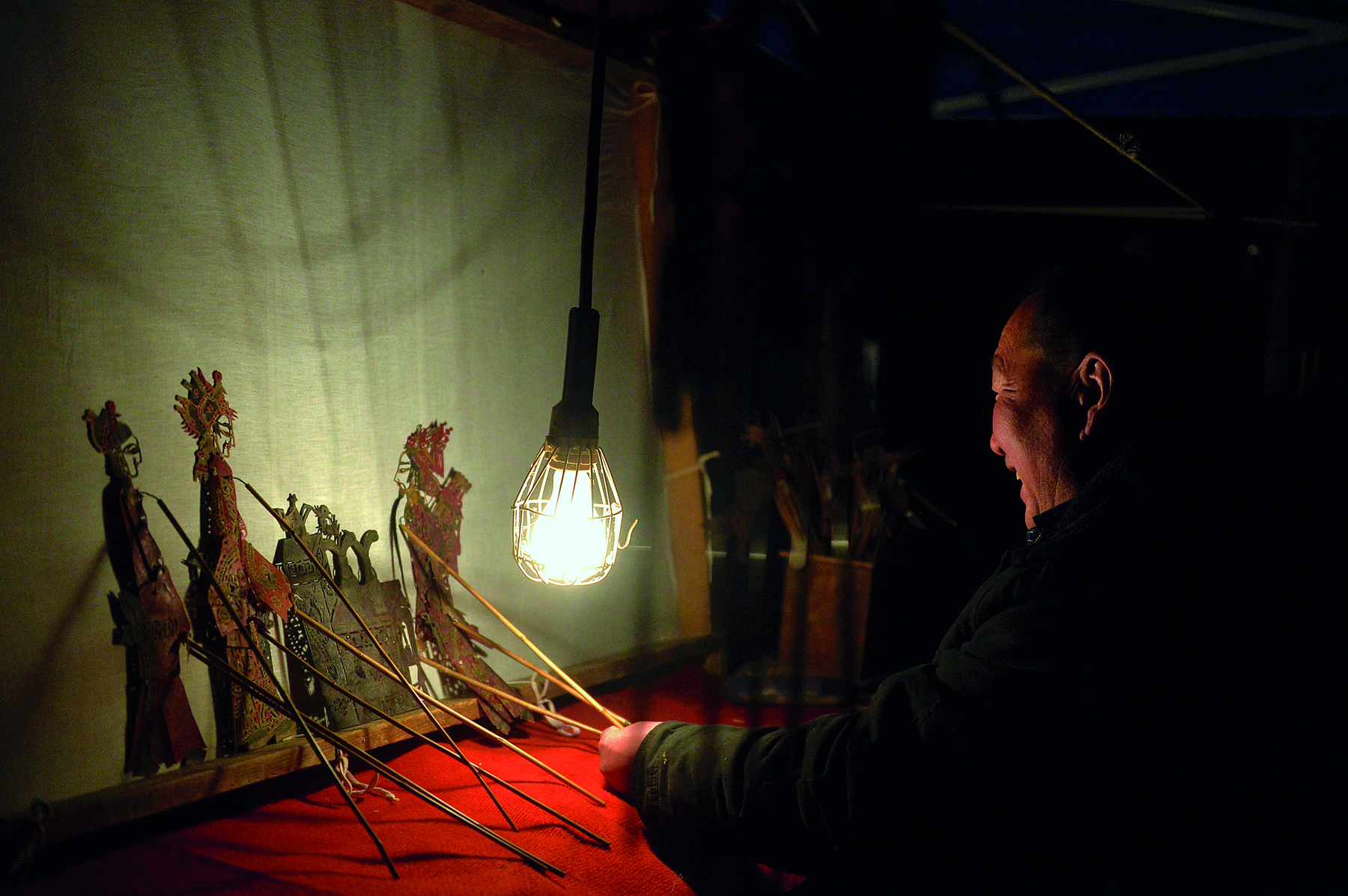 Fotografia. Apresentação de teatro de sombras. Diante de uma lâmpada acesa, um homem está sentado de costas, com o rosto sorridente de perfil, e manipula marionetes em forma de silhuetas diante de uma tela sobre a qual são projetadas as sombras desses personagens, que são observadas pela plateia situada do outro lado da tela, acompanhando o espetáculo.