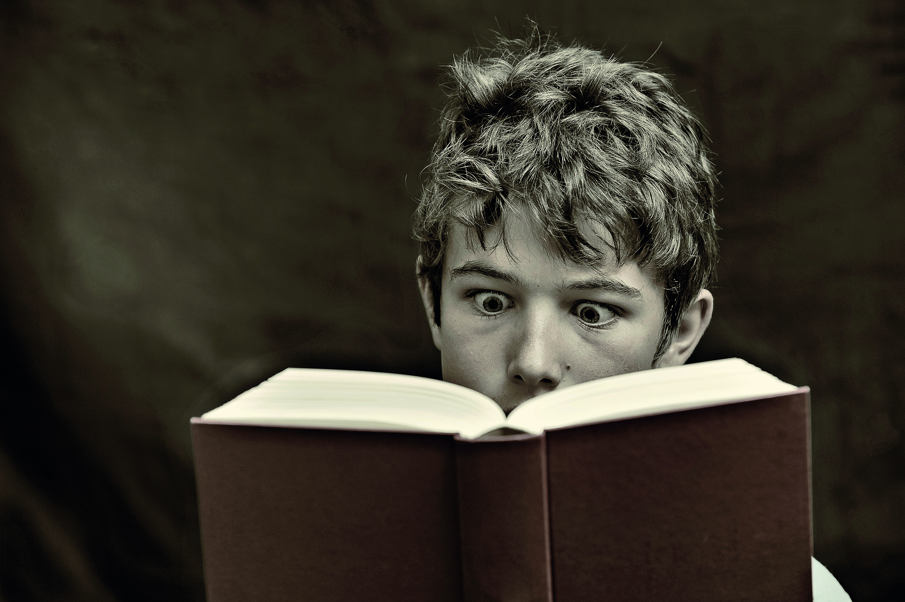 Fotografia em preto e branco. Um jovem de cabelos ligeiramente despenteados segura um livro espesso de capa dura, aberto pela metade, que está diante do seu rosto, cobrindo-o parcialmente. Ele parece absorto pela leitura e tem olhos arregalados e levanta as sobrancelhas, com olhar fixo e vidrado. Ao fundo, um cenário obscurecido.