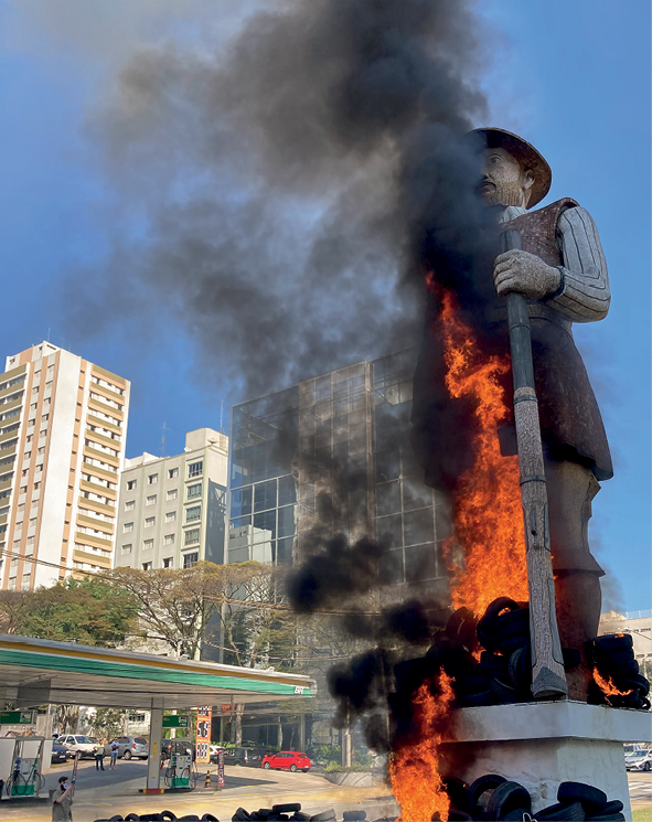 Fotografia. Monumento com estátua de um homem de chapéu calçando botas, com o braço semiflexionado para a frente, segurando uma arma de fogo antiga, que representa a figura de um bandeirante. Há diversos objetos queimando diante do pedestal, e as chamas se elevam até meia altura da estátua, e uma densa fumaça escura a cobre parcialmente. Ao fundo, posto de combustíveis e edifícios sob céu azul sem nuvens.