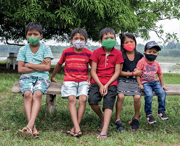 Fotografia. Crianças indígenas vestindo roupas coloridas estão sentadas lado a lado em um banco de madeira. Todas usam máscaras faciais cobrindo o nariz e a boca em um cenário rural, sobre um terreno gramado e com uma árvore de copa extensa atrás deles. Ao fundo há vegetação.