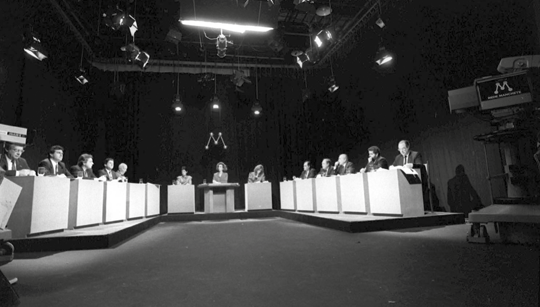 Fotografia em preto e branco. Cenário de uma emissora de tevê no qual ocorre o debate entre candidatos a eleições presidenciais de 1989. Diversos homens de terno e gravata estão sentados em cadeiras diante de tribunas individuais de cor clara e forma cúbica, dispostas sobre um tablado em 'U'. No centro, entre duas filas de candidatos dispostas frente a frente, há três jornalistas, mulheres vestindo tailleur; a mediadora do debate tem uma bancada diante de si. Ao fundo, o símbolo da emissora de tevê que transmite o debate, com a letra 'M'. Acima, diversos refletores de iluminação. À frente, nos cantos da cena, câmeras de tevê.