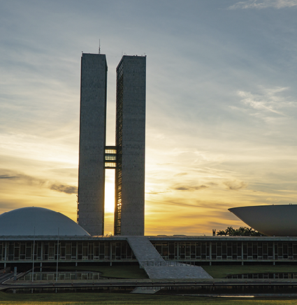 Fotografia. Edifício composto por duas estruturas retangulares idênticas, dispostas lado a lado, unidas por segmentos à meia altura, ladeadas por duas cúpulas brancas circulares, como calotas esféricas, cada uma delas disposta ao lado de uma estrutura retangular. Uma das cúpulas tem a face curva para baixo, e a outra tem a face curva para cima. Abairo desses elementos estruturais há um amplo edifício retangular, sob um céu azul com nuvens brancas e avermelhadas pela luz do Sol que nasce ou se põe.