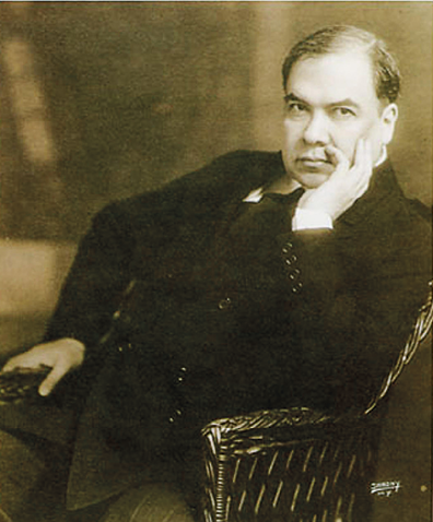 Fotografia em preto e branco. Retrato em busto de um homem com cabelos grisalhos vestindo paletó preto com botões. Ele está sentado em uma cadeira com uma das mãos sobre a perna e a face apoiada na outra mão aberta.