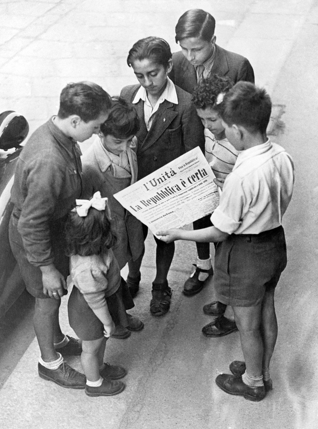 Fotografia em preto e branco. Crianças em uma rua. Um deles, um pré-adolescente vestindo camisa branca e calças curtas escuras está de costas e segura um jornal. Todos são meninos; há apenas uma menina menor que eles, com laço de fita branca nos cabelos. Eles estão ao redor do menino que segura o jornal, interessados na manchete. Os trajes indicam que a cena pertence a um outro tempo, mais antigo.