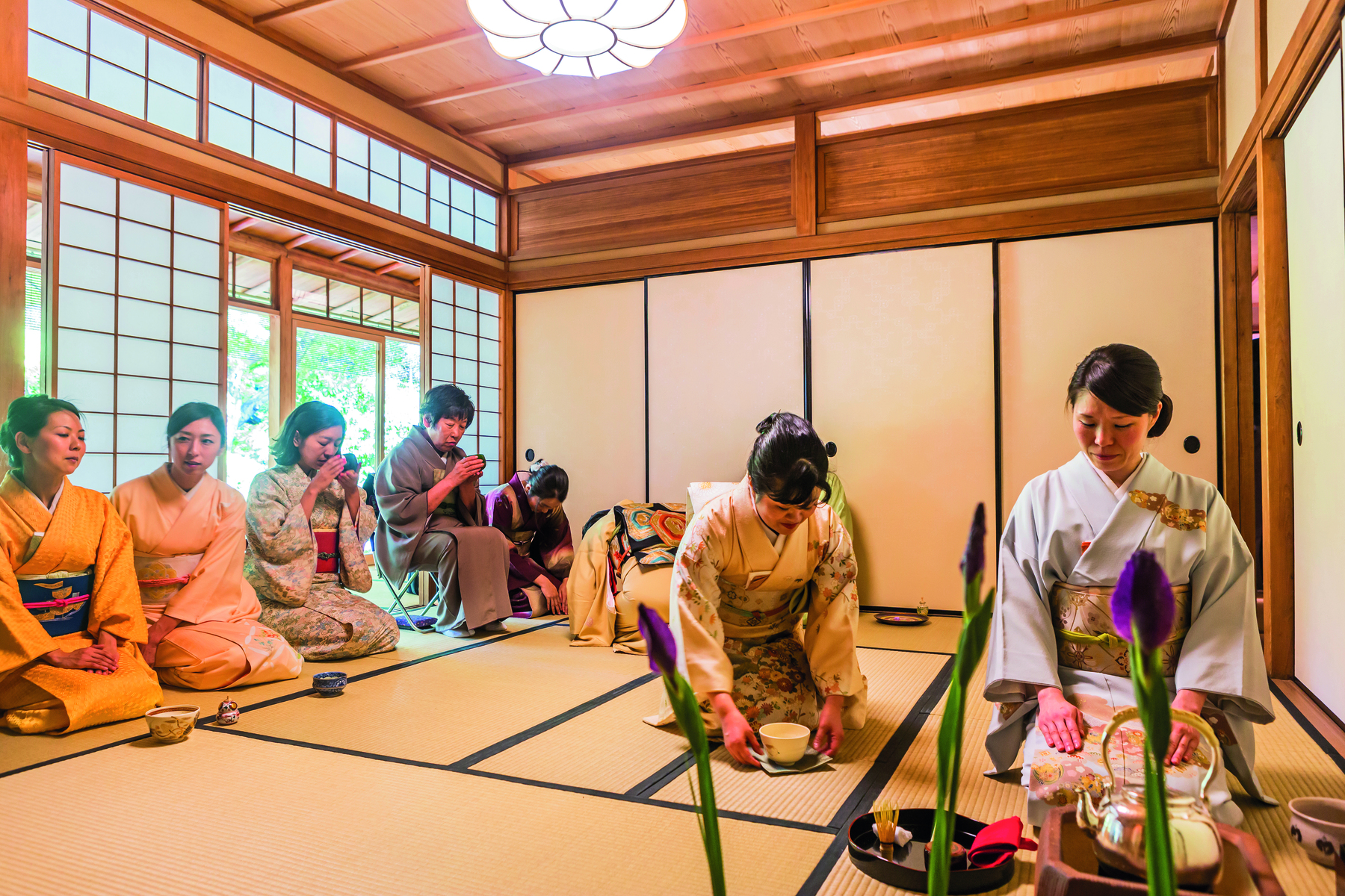 Fotografia. Cerimônia do chá em um aposento tradicional japonês, com estrutura e revestimento de madeira, com portas e janelas revestidas por papel de arroz. Duas mulheres vestindo quimono estão ajoelhadas de frente sobre uma espécie de tatame, diante de um aparato para o preparo de chá. Ao lado, próximas e uma janela, outras mulheres de quimono estão ajoelhadas lado a lado de frente para as primeiras; uma delas está sentada em uma cadeira.