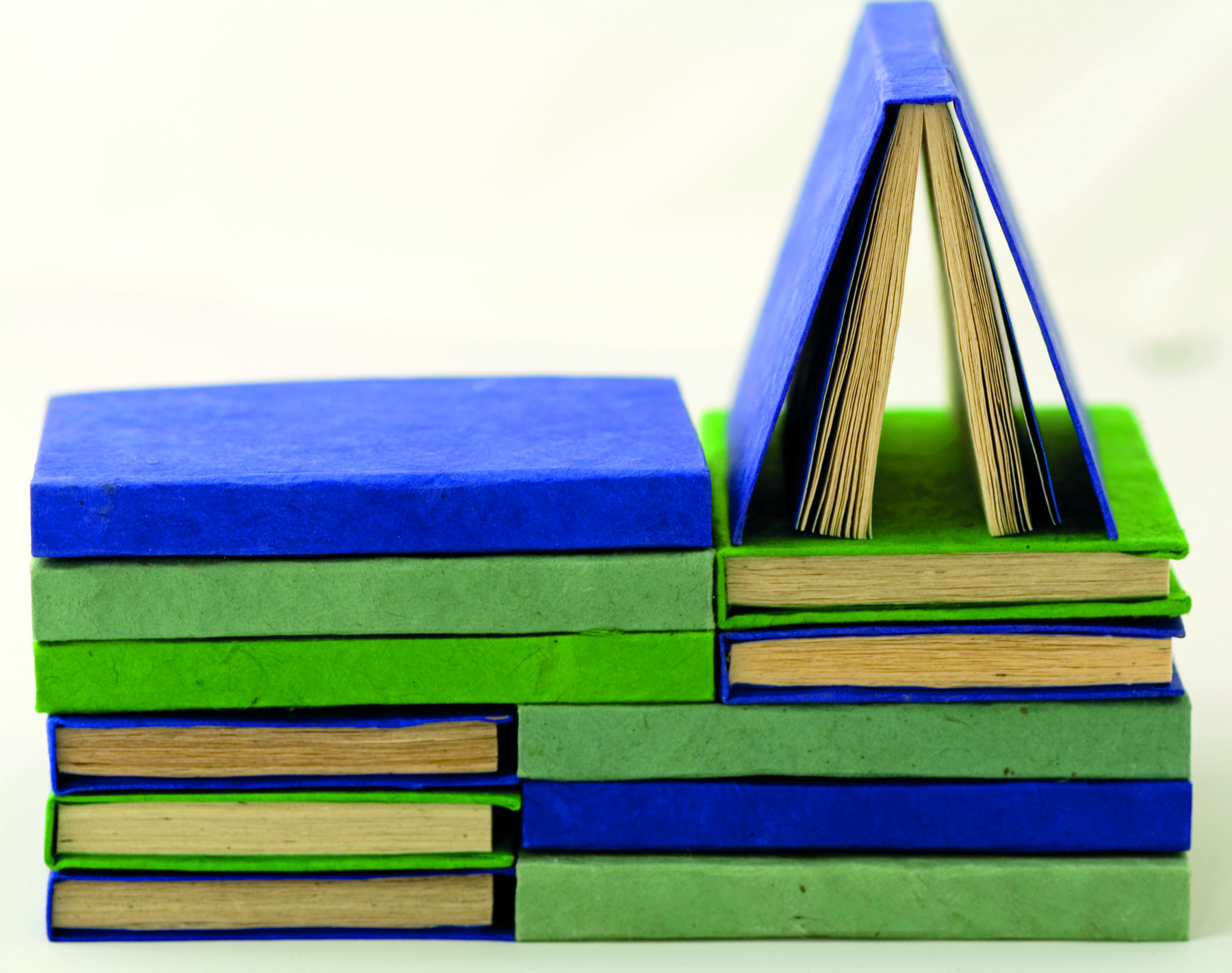 Fotografia. Diversos livros de capas artesanais azuis e verdes empilhados em posições distintas, alguns com a lombada para a frente e outros para o lado esquerdo. À direita, sobre uma das pilhas com a lombada para o lado esquerdo, há um único livro de capa azul semiaberto apoiado sobre um outro livro de capa verde.