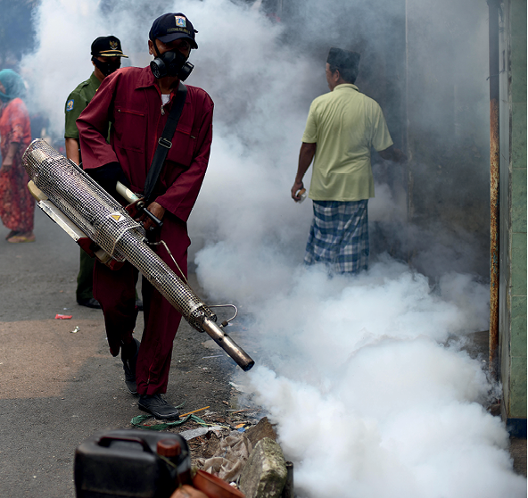 Fotografia. Homem usando macacão alaranjado e máscara contra gases venenosos segura um equipamento de pulverização de inseticida. Há uma densa nuvem branca se estendendo por todo o local onde o inseticida é pulverizado. Em meio a essa nuvem de inseticida há uma pessoa de costas, de camiseta amarela e bermuda xadrez, caminhando no sentido oposto, com a cabeça ligeiramente voltada para o lado, de modo a revelar que não usa máscara alguma.