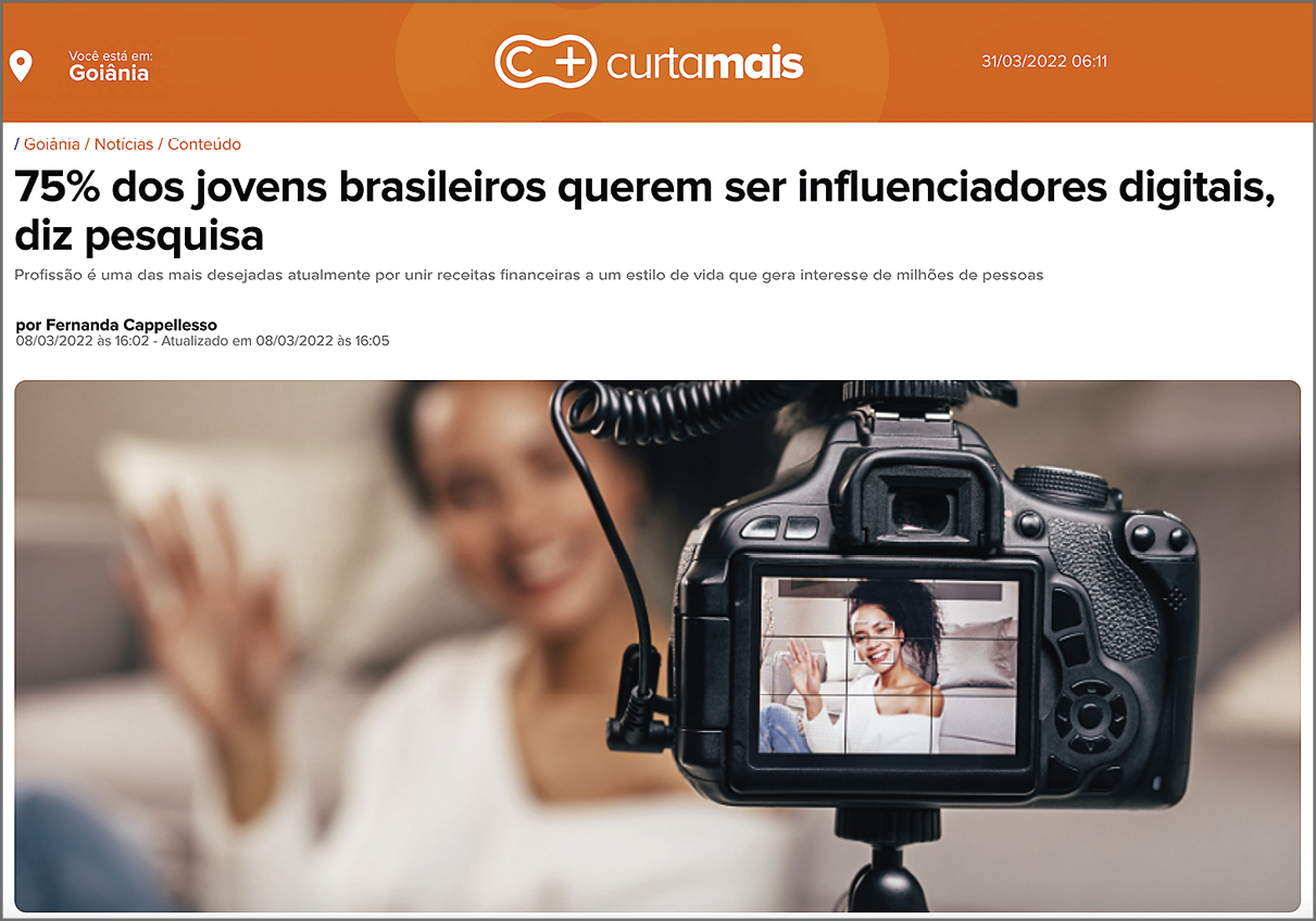 Fotografia. Reprodução de página de internet. Acima, o título da página web: 'C+ curta mais'. Abaixo, a manchete: '75% dos jovens brasileiros querem ser influenciadores digitais, diz pesquisa' e o nome da autora da matéria: 'por Fernanda Cappellesso'. Abaixo, fotografia de uma câmera fotográfica apoiada na extremidade superior de um tripé, cuja tela enquadra uma jovem/adolescente sorridente de cabelos crespos presos para o lado, com um braço flexionado para cima e a mão espalmada. Ela veste uma blusa branca que revela o pescoço, parte do tronco e um dos ombros. Ao fundo, a jovem/adolescente de cabelos presos enquadrada pela câmera.