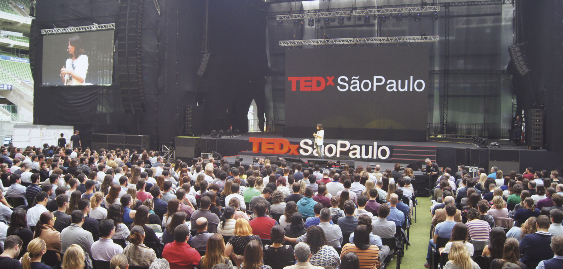 Fotografia. Retrato da apresentação de uma palestra no centro do palco de um auditório. A palestrante está em pé, mas a distância não possibilita perceber outros detalhes dela, pois a imagem enfatiza a plateia, praticamente lotada que está diante dela: inúmeras pessoas sentadas, de costas. No palco, atrás da palestrante, há grandes letras tridimensionais formando o título do evento e o local: 'TEDx' em vermelho, e 'São Paulo', em branco. Ao fundo, uma tela cinza-escura, na qual são projetadas estas mesmas palavras, e ao lado, uma outra tela exibindo a palestrante en close.