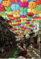 Imagem: Fotografia. Trecho de uma rua com construções assobradadas e onde pessoas estão sentadas em cadeiras, outras caminham e há um corredor central onde há sombrinhas abertas. Todo espaço é coberto por uma composição de sombrinhas abertas coloridas que formam um teto.  Fim da imagem.