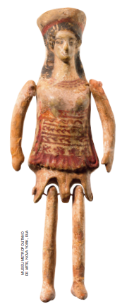 Imagem: Fotografia. Uma boneca de uma mulher de pé com braços e pernas longas, detalhes pintados sobre a vestimenta, membros unidos com arame, cabelo longo e um acessório sobre a cabeça.  Fim da imagem.