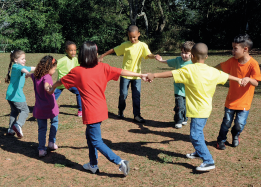 Imagem: Fotografia. Em uma área gramada, oito crianças de pé estão de mãos dadas formando uma roda. Fim da imagem.