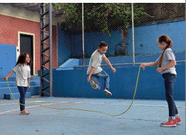 Imagem: Fotografia. No pátio de uma escola, três crianças uniformizadas brincam de pular corda. Fim da imagem.