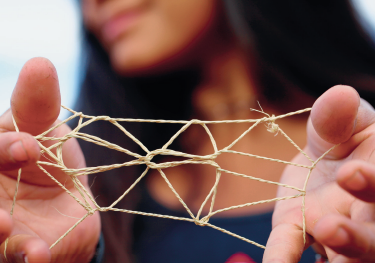 Imagem: Fotografia. Destaque das mãos espalmadas em paralelo de uma menina que está com um cordão de fibra entrelaçado com nós entre os polegares e dedos mindinhos.  Fim da imagem.