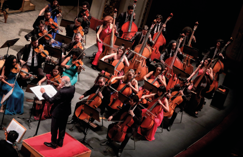 Imagem: Fotografia. Em um espaço fechado, vista superior de um grupo de pessoas tocando instrumentos, como violinos e violoncelos, em uma orquestra sinfônica. Eles formam uma composição em fileiras, alguns estão sentados e outros de pé. Diante de todos, há um senhor de terno preto sobre uma estrutura no alto. Ele segura uma baqueta e está diante de partituras.  Fim da imagem.