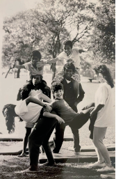 Imagem: Fotografia em preto e branco. Um grupo de oito crianças em uma área verde com árvores. Em primeiro plano, um menino é segurado pelos braços e pelas pernas por duas meninas. Ao fundo, dois meninos sustentam outros dois colegas sentados em seus ombros.  Fim da imagem.